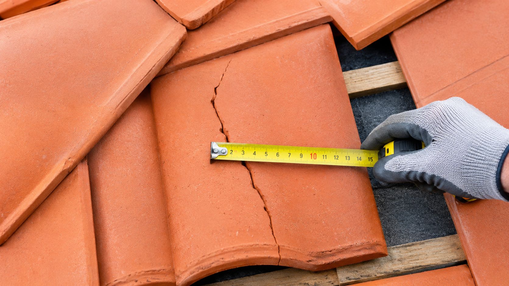A gloved hand uses a yellow tape measure to gauge the size of a cracked terracotta roof tile.
